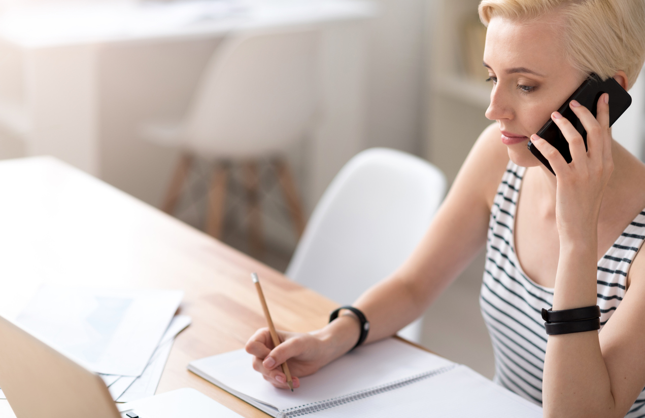 Woman taking notes on a notepad while talking on the phone and looking at the laptop. Looking to acquire top talent.