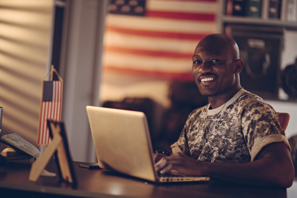 Picture of a young smiling african american soldier who is sitting by a desk using a laptop. On the desk is also a small american flag, a photo frame and in the background a bookshelf and a nice dimmed light sipping threw a window from the left. The soldier is looking in to the camera with a big smile.