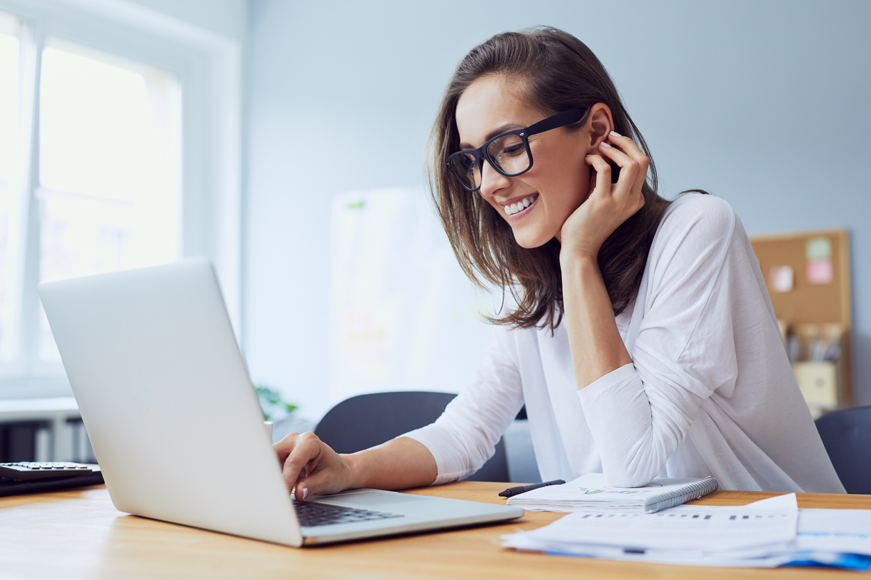 Portrait of beautiful cheerful young businesswoman working on laptop and laughing in home office. Employee engagement concept.