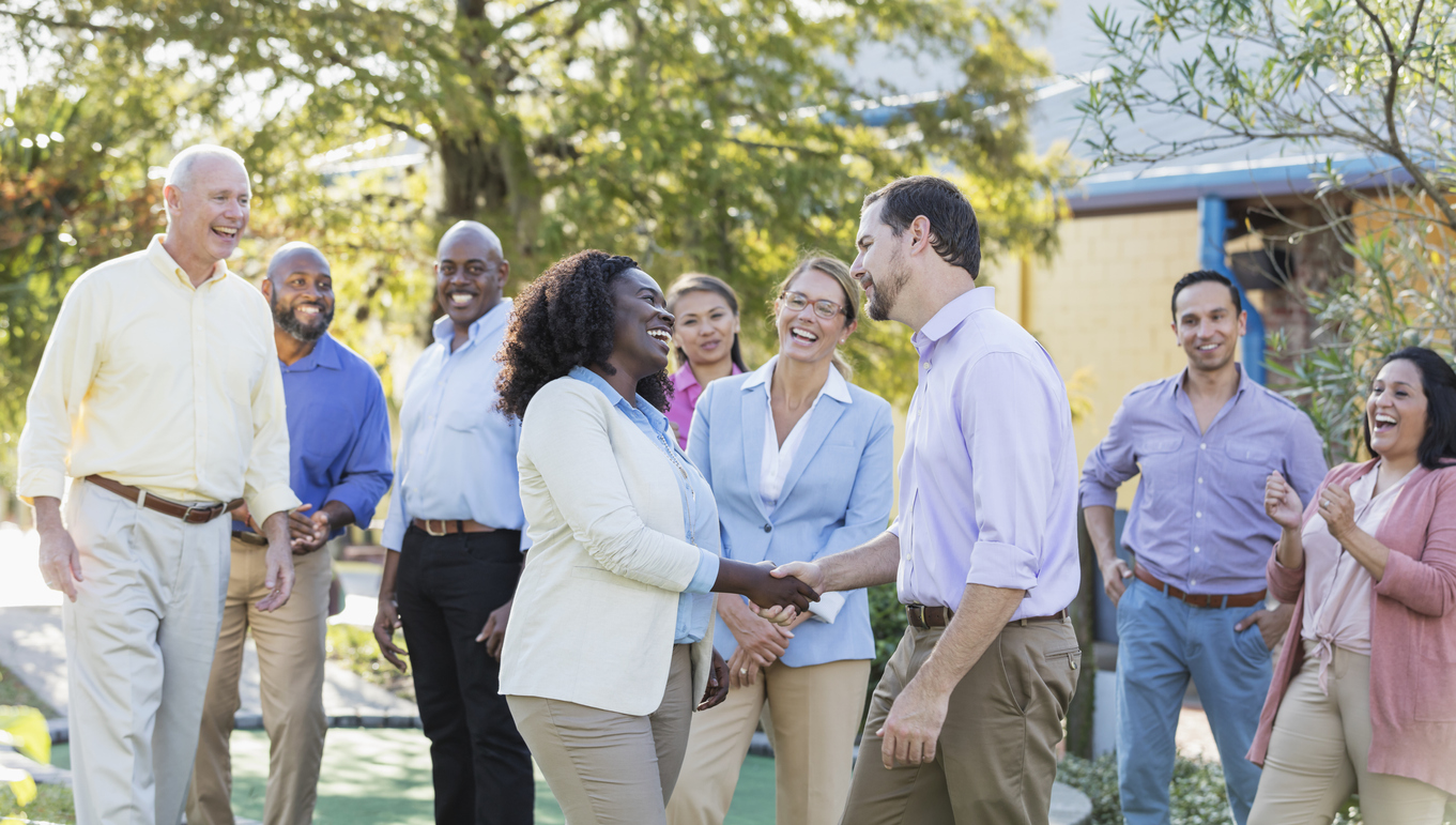 A multi-ethnic group of nine business people participating in corporate team building and bonding activities, standing outdoors. The coworkers range in age from 20s to 60s. The focus is on the young African-American woman, in her 20s, and the mid adult man, in his 30s, standing with her in the foreground. They are shaking hands as their colleagues watch.