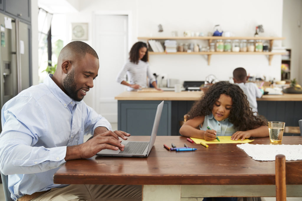 Young black family busy working in their kitchen. Father seeking jobs with health benefits on his laptop.