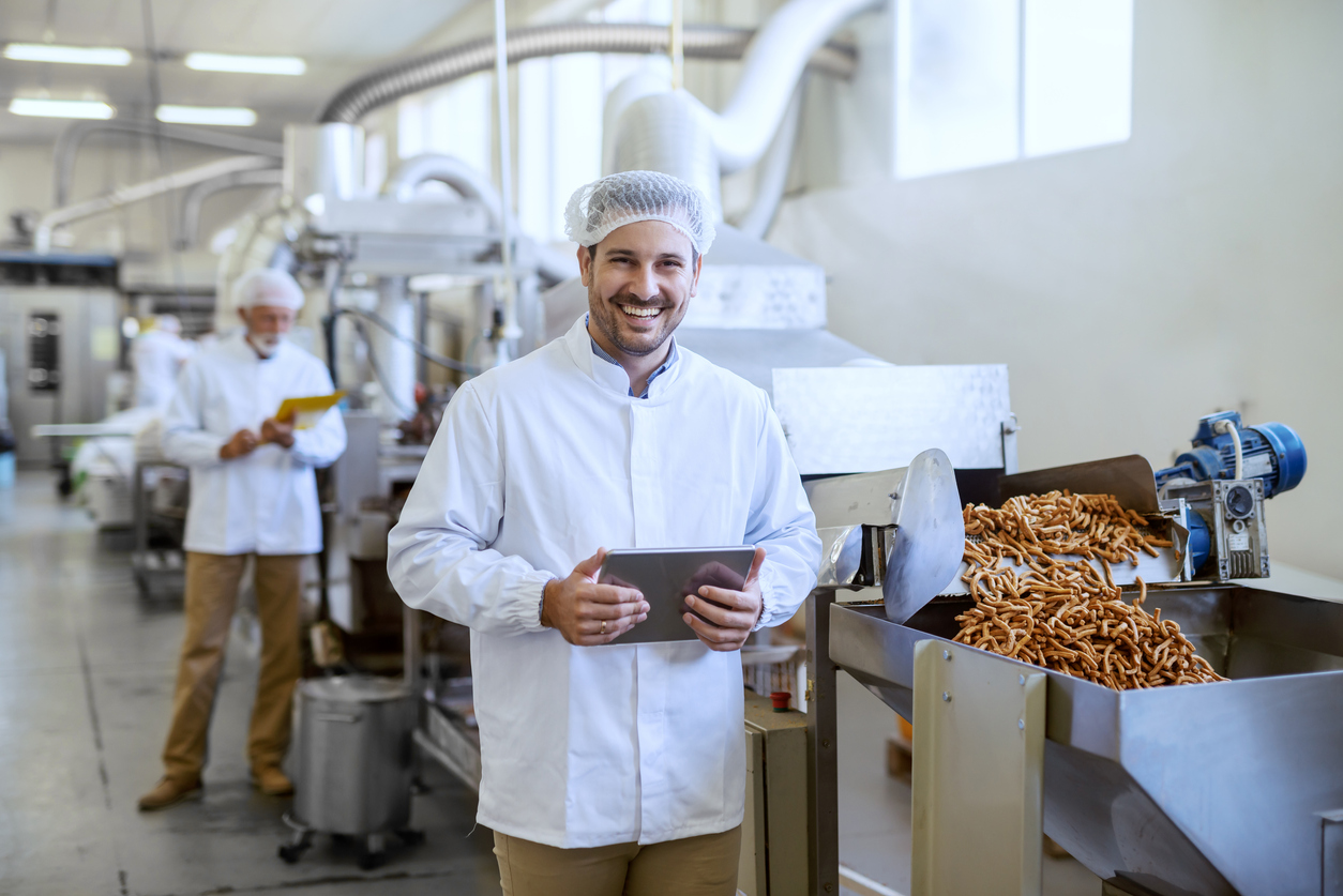 Young smiling manager in sterile uniform holding tablet and looking at camera while standing in a manufacturing food factory.