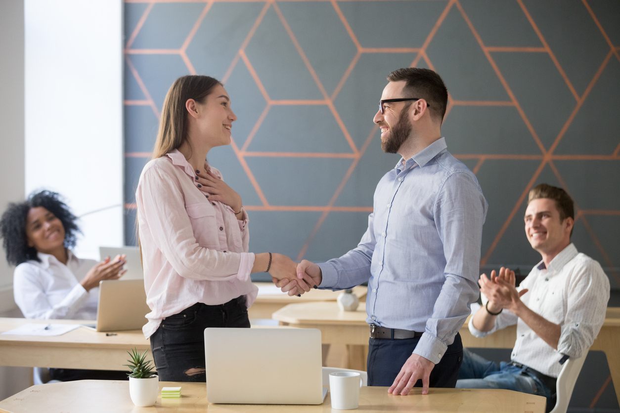 Male boss or team leader shaking hand of female temporary employee congratulating with hiring her for a full-time position, appreciating for good work result while business team applauding, recognition concept