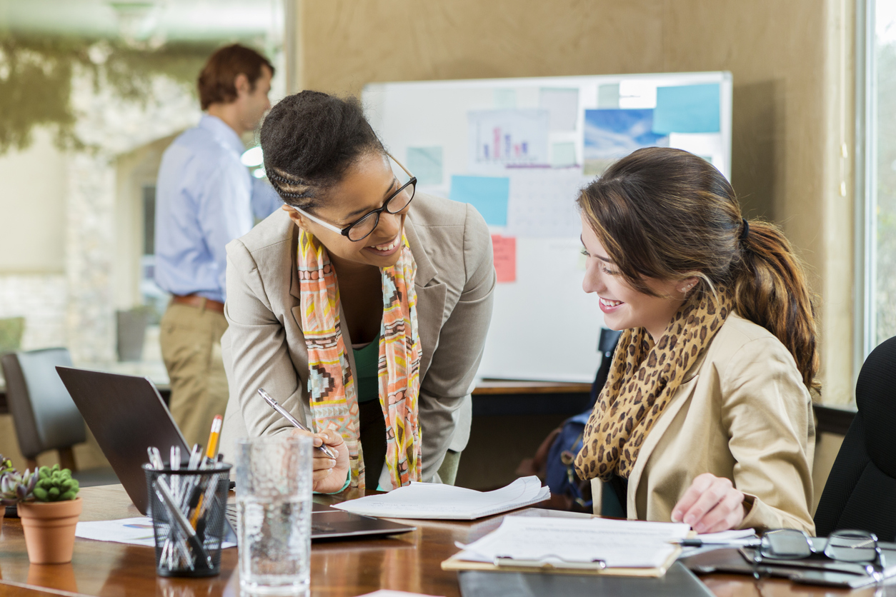 Executive and Executive assistant discuss something in the office. They are reviewing documents at the Caucasian woman's desk. A man is working in the background.