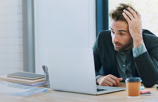 A man is looking at his laptop overwhelmed, holding his head in his hand.