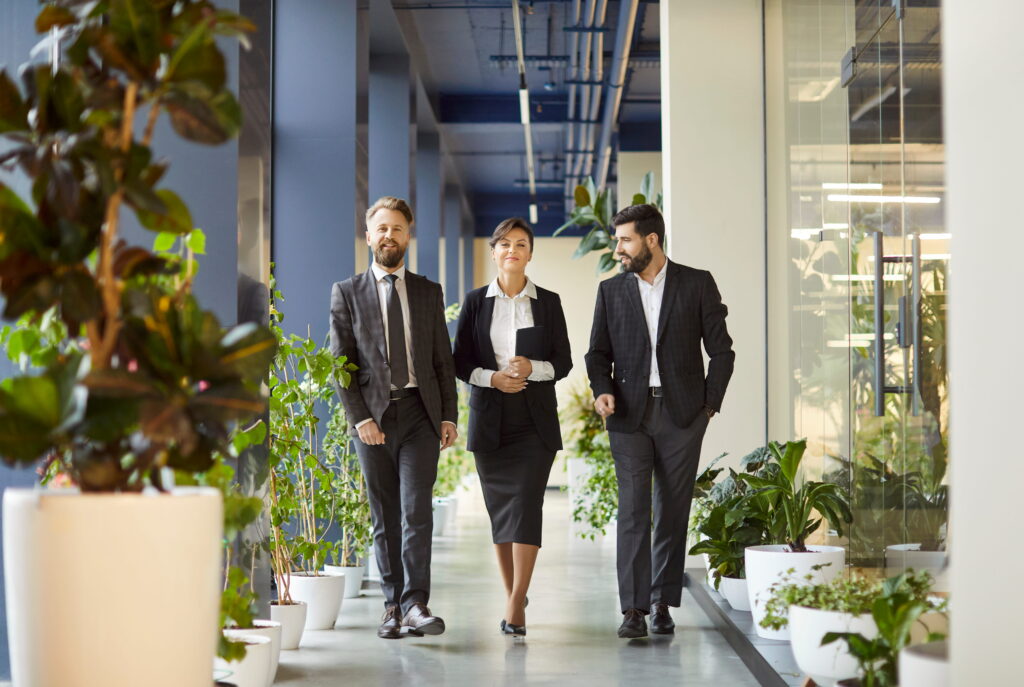 Three business professionals in suits walking through modern office corridor surrounded by green plants. Confident colleagues discussing work, sharing ideas and collaborating in corporate environment. Downloaded File #: 1368716672 Three business professionals in suits walking through modern office corridor surrounded